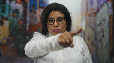 Şehir sokaklarında kendine güveni tam bir kadın, güçlü, iddialı bir tavır sergiliyor, el hareketi, kalın çerçeveli gözlükler ve ciddi bir ifadeyle kendini gösteriyor..