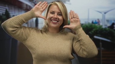 Gülümseyen sarışın kadın, aydınlık modern havaalanı terminalinde yüzün yanındaki ellerini çerçeveliyor; mutluluk.