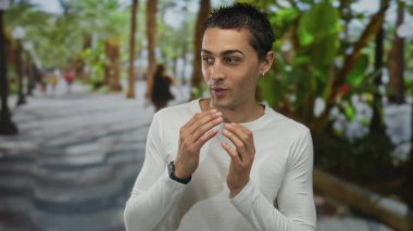 Genç İspanyol asıllı bir adam güneşli parkta palmiye ağaçları ve arka planda bulanık insanlarla jest yaparak rahat ve rahat bir atmosfer yayıyor..