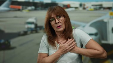 Üst düzey İspanyol kadın havaalanı terminalinde göğüs ağrısı yaşıyor. Arka planda uçaklar seyahat stresi ya da acil bir sağlık sorunu olduğunu gösteriyor.