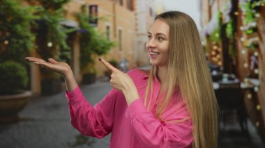 Pembe gömlekli genç bir kadın, güzel bir gökyüzü altında canlı bir açık hava yemek deneyimini somutlaştıran, restoran atmosferli büyüleyici bir terasta avucunu gösteriyor..