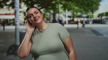 Şehir caddesinde ağaçlarla gülümseyen ve arka planda insanlar olan bir kadın, canlı bir açık hava sahnesini şehir ortamında yakalıyor..