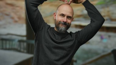 Kel ve sakallı bir adam, arka planda okyanusla birlikte sahilde gülümsüyor, açık havada günlük giysiler giyiyor ve güneşli bir günün tadını çıkarıyor..