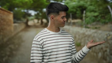 Çizgili tişörtlü ve elini açık bir parkta dikilen genç bir adam. Arka planda taştan bir yol ve yemyeşil bir zemin var. Neşeli görünüyor..