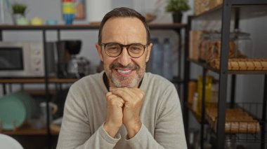 Ofis ortamında gülümseyen bir adam, modern dekorlar, şişeler ve tabaklarla çevrili, sıcaklığını ve özgüvenini sergiliyor, profesyonel, sakin bir atmosferi gösteriyor..