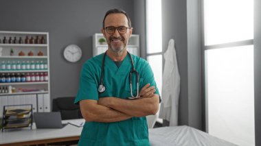 Olgun İspanyol bir adam, klinik odasında güvenle ayakta duruyor. Kollarını kavuşturup kameraya gülümsüyor ve kapalı alanda profesyonel bir sağlık hizmeti ortamı yaratıyor.