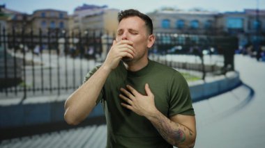 Şehir dışında hapşıran genç bir adam yeşil gömlekli ve dövmeli bir adam. Gün boyunca bulanık şehir arka planına karşı görünür..