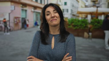 Orta yaşlı, kollarını kavuşturmuş İspanyol kadın gün ışığında şehir caddesinde duruyor..