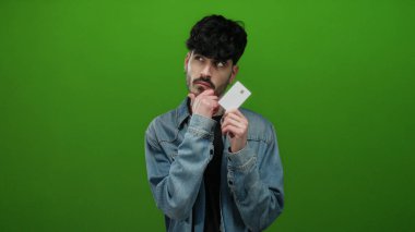Genç adam yeşil bir arka plana karşı duruyor, elinde bir kart ve kot ceket tutuyor, düşünceli bir şekilde çenesi ve gözleri kapalı poz veriyor..