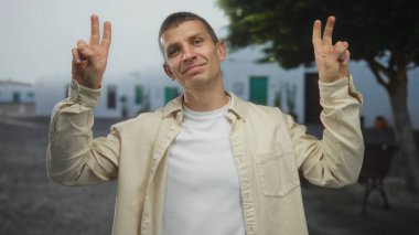 Açık bir sokakta bulanık beyaz binalar ve ağaçlarla dikilen, günlük gömlekler giyen ve pozitif ifade gösteren bir adam..