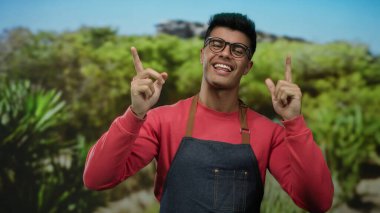Önlüklü ve gözlüklü genç adam gülümsüyor ve canlı yeşil bir açık hava parkında dikiliyor..