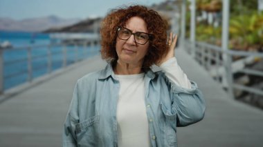 Arka planda dağlarla ve okyanuslarla dolu deniz kenarında el kol hareketleri yapan, gözlük takan ve günlük giysiler giyen, açık havada duygularını gösteren bir kadın..