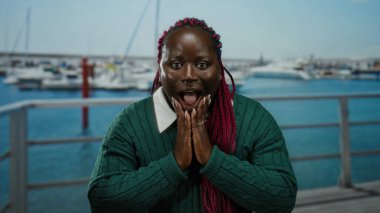 Saçları örgülü bir kadın, arka planda teknelerle dolu bir limanda, canlı duygular sergileyen açık havada bir anı yakalayarak şaşkınlığını dile getiriyor..