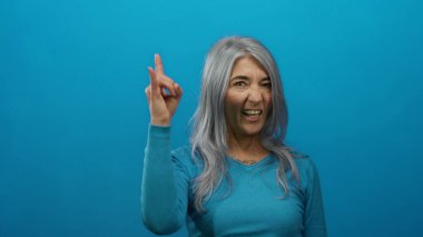 Gri saçlı yaşlı kadın, izole edilmiş mavi bir arka plan duvarına karşı duran, güven ve enerjiyi yansıtan bir kaya hareketi yapıyor..