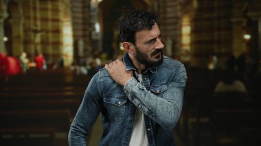 Kot ceketli, sakallı bir İspanyol Katolik kilisesinde duruyor ve omzunu tutarken rahatsız olduğunu söylüyor..