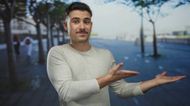 Latin kökenli genç bir adam şehir dışındaki bir sokak manzarasına el uzatıyor. Arka planda ağaçlar olan bir şehir manzarası sunuyor. Geniş ve misafirperver bir ortam yaratıyor..