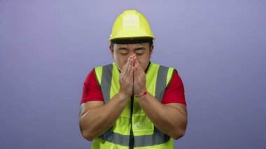 Kask ve güvenlik yeleği giyen genç adam, izole edilmiş mor bir arka plan önünde soğuk görünüyor. Rahatsızlık ve ürperti hislerini iletiyor..