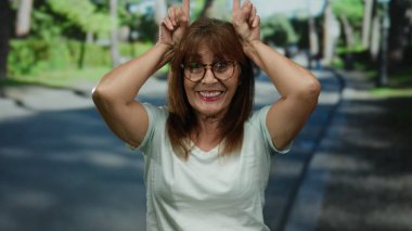 Kıdemli İspanyol kadın neşeli bir gülümseme ve gözlüklerle güneşli bir günde neşe içinde parkta poz veriyor..
