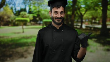 Siyah üniformalı ve eldivenli İspanyol şef yeşil bir parkta ağaçlarla baş parmak işareti yapıyor, kameraya güvenle gülümsüyor..