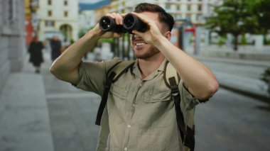 Şehrin hareketli sokaklarında dürbün kullanan sakallı genç bir adam, açık bir gökyüzünün altında sırt çantasıyla şehir ortamını keşfeden meraklı bir turisti gösteriyor..