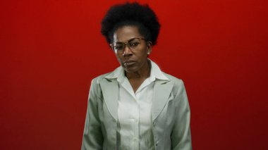 Gözlüklü bir kadın, canlı kırmızı bir arka planda, beyaz bir bluz ve takım elbise giyiyor, düşünceli bir şekilde düşünüyor ve kendine güveniyor..