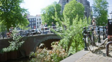 Bulanık insanlar güneşli, dağılmış bir sahnede bisiklet, ağaç ve tarihi mimari ile pitoresk bir kanal köprüsü boyunca yürüyorlar..