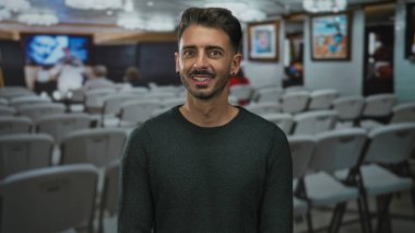 Yeşil kazak giyen bir adam binanın içindeki çerçeveli resme gülümseyerek parmak işareti yapıyor; merak neşe kaynağı..