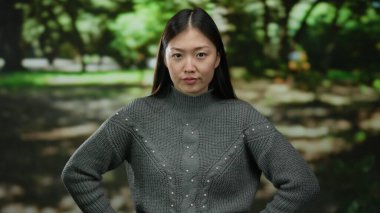 Gri kazaklı bir kadın, gün içinde açık havada bulanık yeşil arka planlı bir parkta güvenle duruyor. Güçlü ve kararlı bir ifadeyi somutlaştırıyor..