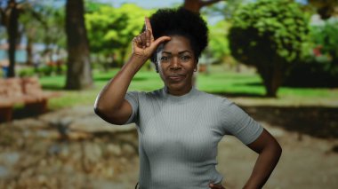 Parkta kendine güveni olan bir kadın, arka planda ağaçlar ve yeşilliklerle ezikçe bir jest yapıyor mizah ve basitliği resmediyor..