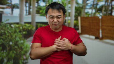 Açık havada göğsünü tutan kırmızı gömlekli genç adam bulanık şehir geçmişiyle çevrili rahatsızlığını dile getiriyor. Açık hava ortamında kalp ağrısı ya da sağlık sorunu olduğunu gösteriyor..