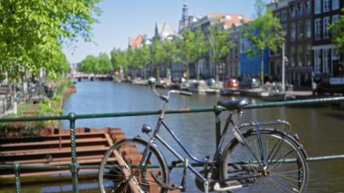 Bisiklet, açık bir günde arka planda bulanık bir kanalı ve tarihi evleri olan köprü parmaklıklarında dinleniyor. Hollanda kentsel manzarasının özünü yakalıyor..