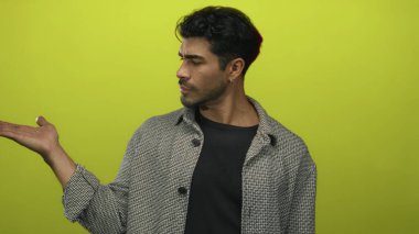 Sarı ceketli İspanyol genç adam, sarı stüdyoda avuç içini ve parmağını açık tutuyor; belirsizlik..