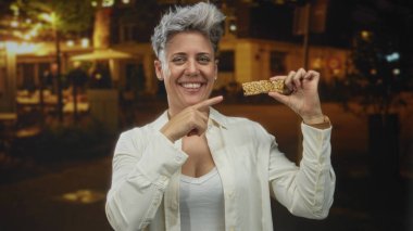 Gece, elinde granola çubuğuyla gülümseyen ve kentin caddesinde atıştırmak için parmağını gösteren bir kadın. Mutluluk..