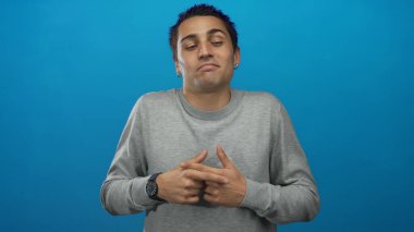 Genç İspanyol adam belirsiz bir ifadeyle gri süveter giyerek izole edilmiş mavi arka plana omuz silker.