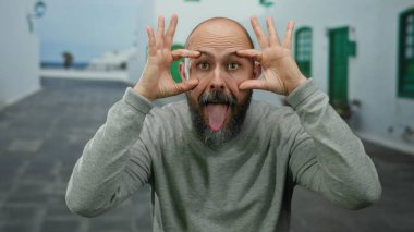 Sakallı kel bir adam, şehir merkezinde komik suratlar yapıyor. Açık havada elinde dövmesi olan gri bir kazak giyiyor..