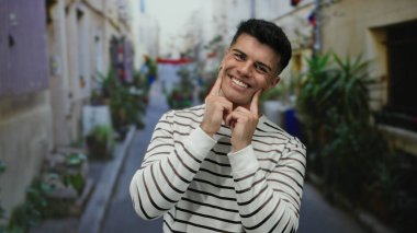 Neşeli bir şehir sokağında, çizgili bir tişört giymiş, güneşli bir günde, yemyeşil bir sokaktaki çiçeklerle çevrili, kendine güvenen genç bir adam..