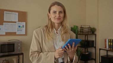 Ofiste elinde kendinden emin bir gülümsemeyle tablet tutan kadın, duvarında çizelgeleri ve şık iç dekorasyonu olan modern işyerini gösteriyor, profesyonelliği ve nişanı iletiyor..