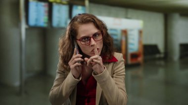 Havaalanı terminalinde telefonla konuşan, kırmızı gözlüklü ve bej ceketli bir kadın bilgi ekranlarının ve satış makinesinin bulanık arka planına karşı konuşuyor..