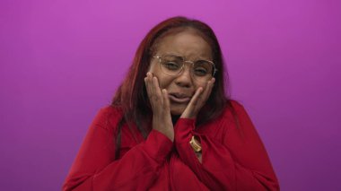 Stüdyoda eli göğsünde kırmızı bluz giyen ve yuvarlak gözlükler takan mor arka planı olan bir kadın..