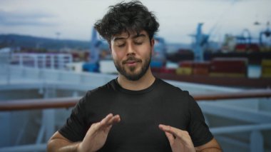Genç İspanyol bir adam sakin bir şekilde meditasyon yapıyor. Gözleri kapalı, arka planda gemileri ve botları olan bir limanda. Denizcilik ortamında sükuneti somutlaştırıyor..
