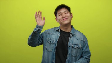 Sarı arka planda gülümseyen ve kot ceketli genç Çinli adam dostça bir selamlama ve olumlu bir ruh hali içinde.
