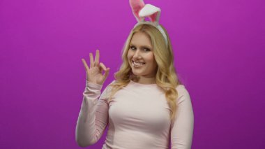 Tavşan kulağı takan ve parlak pembe arka plana karşı iyi bir hareket yapan, neşeli davranışlar sergileyen, sarışın ve genç görünümlü neşeli ifadeler sergileyen bir kadın..