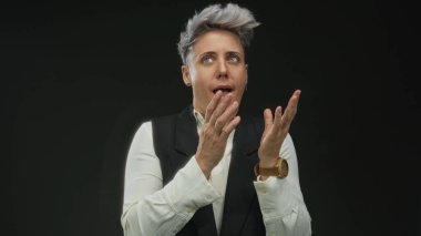 Siyah yelek ve beyaz gömlek giyen kısa gri saçlı kadın siyah stüdyoda parmaklarıyla dudaklara dokunuyor..