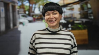 Kısa saçlı bir kadın açık hava restoranının terasında komik surat yapıyor. Geçmişi bulanık. Çizgili süveter ve küpeler takıyor..