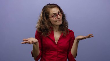 Kırmızı gözlüklü ve gömlekli, mor bir arkaplan karşısında el kol hareketi yapan kadın, belirsizliği ifade eden sakin bir yüz ifadesiyle kafa karışıklığı gösteriyor..