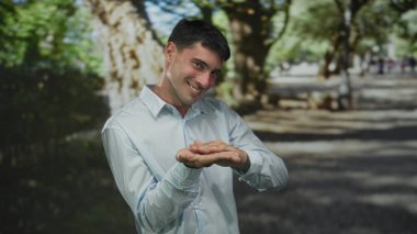 Açık mavi gömlekli genç İspanyol adam, güneş ışığında yeşil ormanda para için parmaklarını ovuşturuyor..