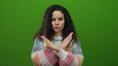 Kıvırcık saçlı, renkli kazaklı bir kadın kollarını yeşil arka planda kavuşturmuş.