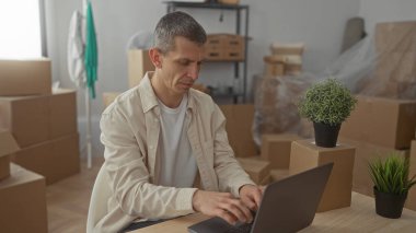 Dizüstü bilgisayarda çalışan sıradan giysili bir adam. Etrafı karton kutular ve bitkilerle çevrili. Yeni apartman dairesinde..