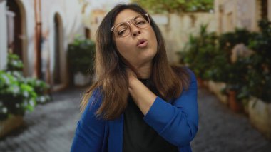 Güneş ışığıyla aydınlanan eski şehir sokağında, saksıların ve yıpranmış taş duvarların arasında bir kadın omuz ve irin tutuyor..