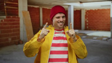 İnşaat alanında sarı yağmurluk ve kırmızı bere giyen kendine güvenen, çizgili gömlekli çekici bir adam..